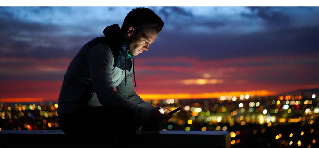 Young man at night sitting on a wall looking at his phone with city lights in the background