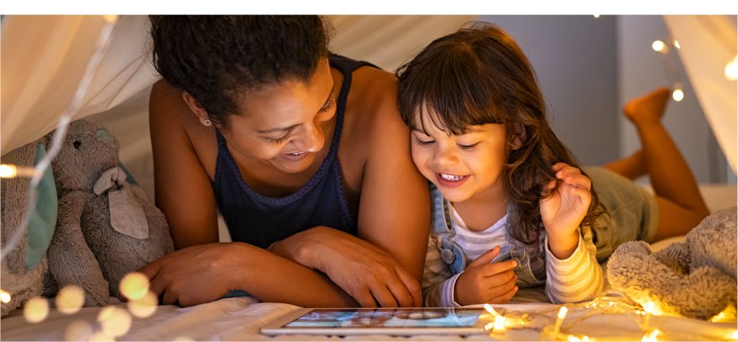Happy woman with child inside a tent with fairy lights looking at a tablet.