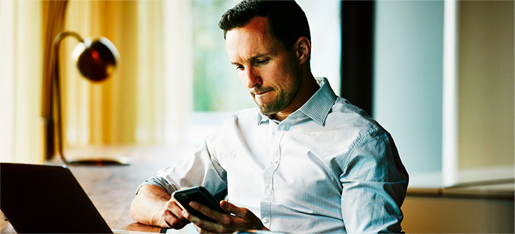 Man sitting at table with laptop, whilst scrolling through his phone.