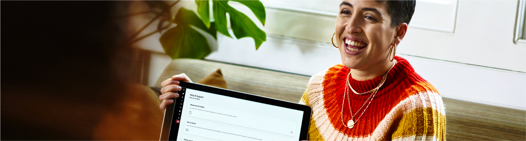 Female support adviser with multi-coloured sweater holds a laptop whilst chatting with customer in a bright office