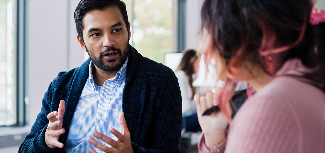 Two people engaged in conversation in a modern office setting, with large windows allowing natural light to fill the space