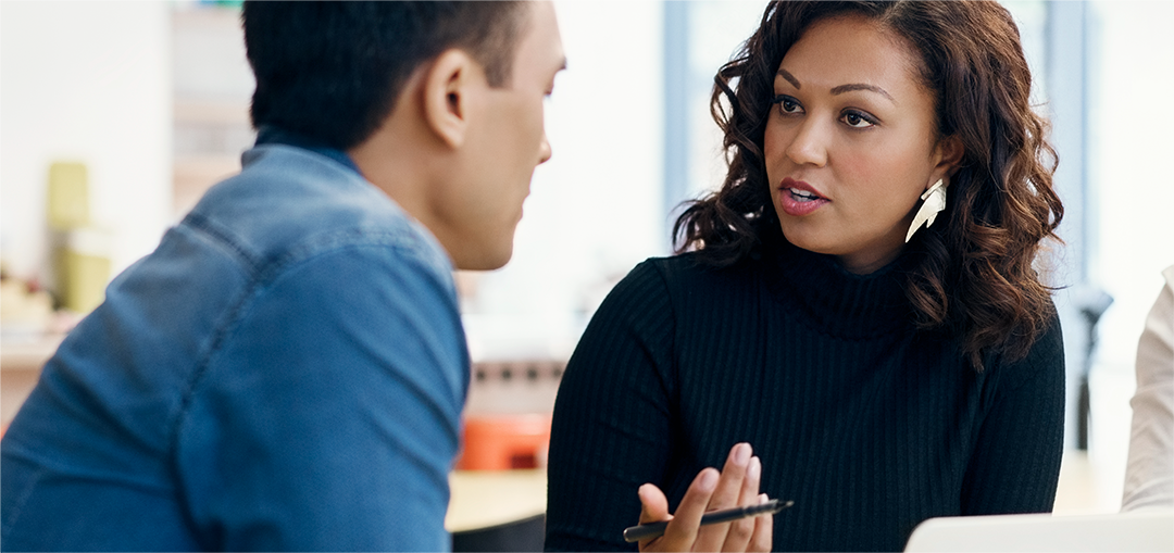 Two people engaged in a conversation, with one wearing a blue shirt and the other in a dark sweater, in a softly lit indoor setting