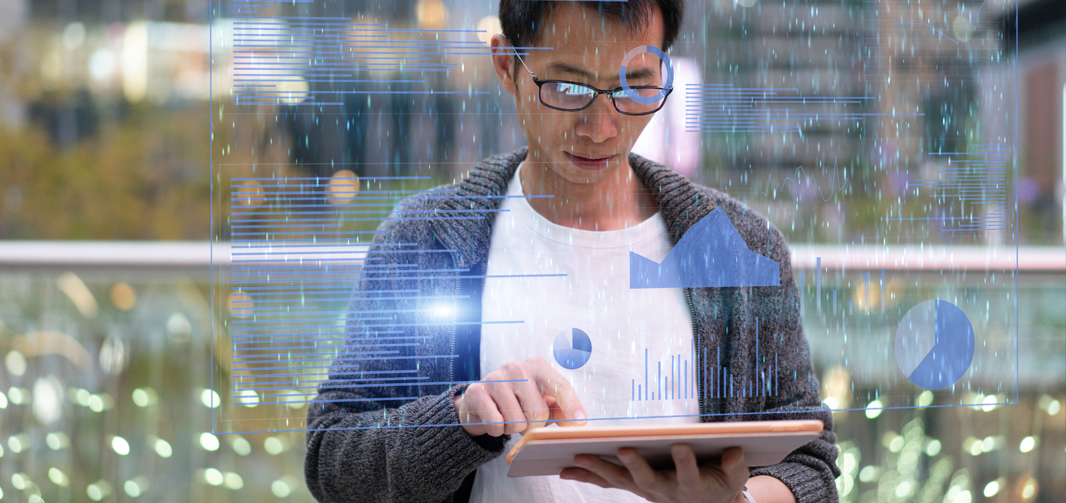 Man standing looking at a tablet with holographic charts and graphs floating in front of him.