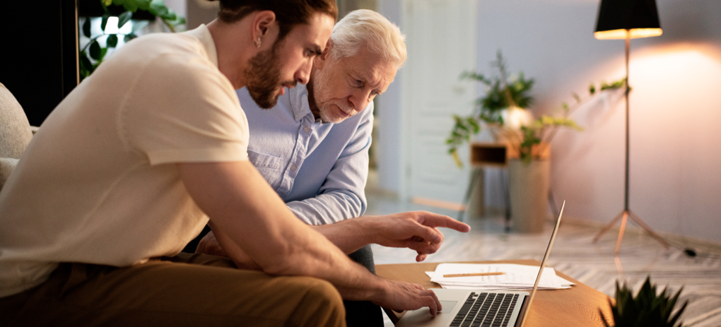 Young man with beard and a white shirt discusses finances on a laptop with an older man with grey hair and blue shirt