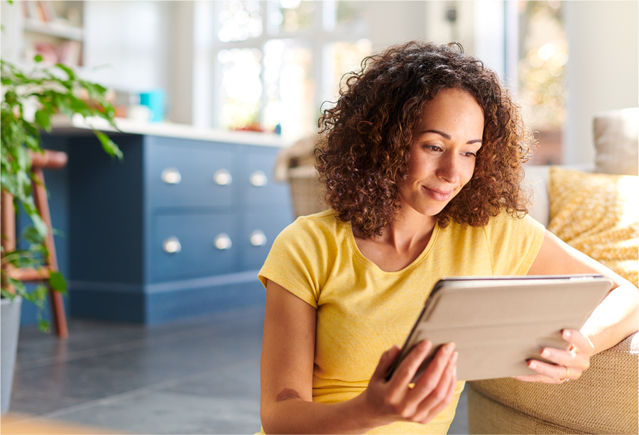 Young woman with long curly brown hair and wearing a yellow top sits on the floor in her house whilst looking at a tablet weighing up her investment options