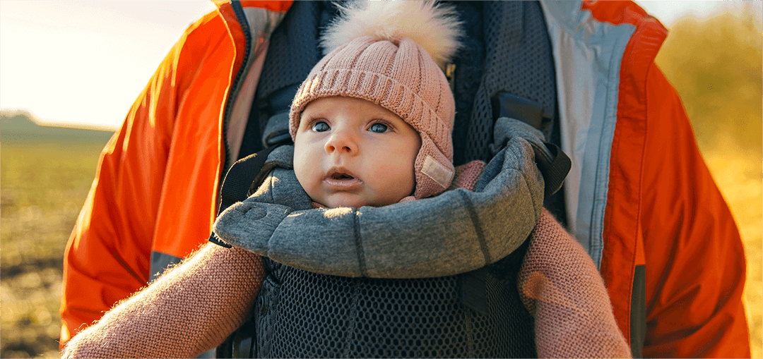 A close-up of a person wearing an orange jacket, holding a baby in a grey carrier, with soft sunlight illuminating a grassy field