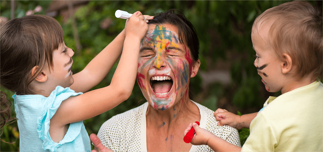 Two children playfully draw on an adult's head with colourful markers, surrounded by lush greenery in a playful outdoor setting