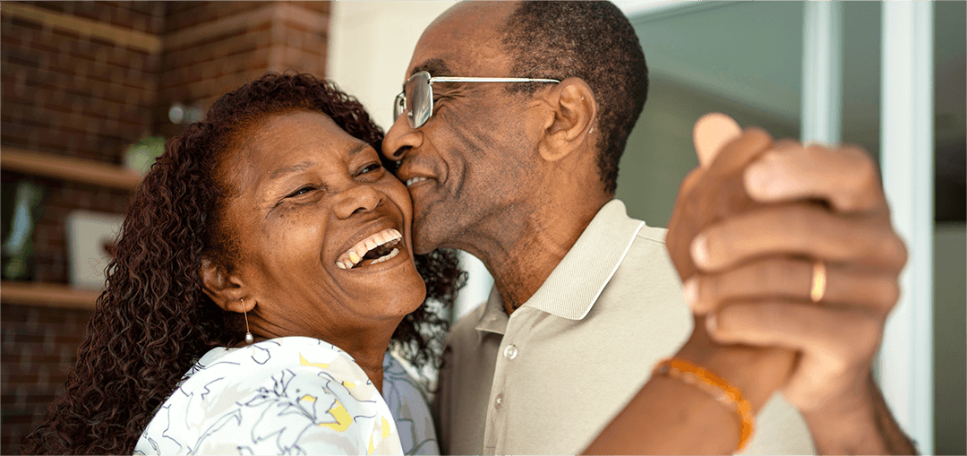 A couple joyfully holds hands, dancing closely in a cosy indoor setting with warm lighting and a brick wall in the background
