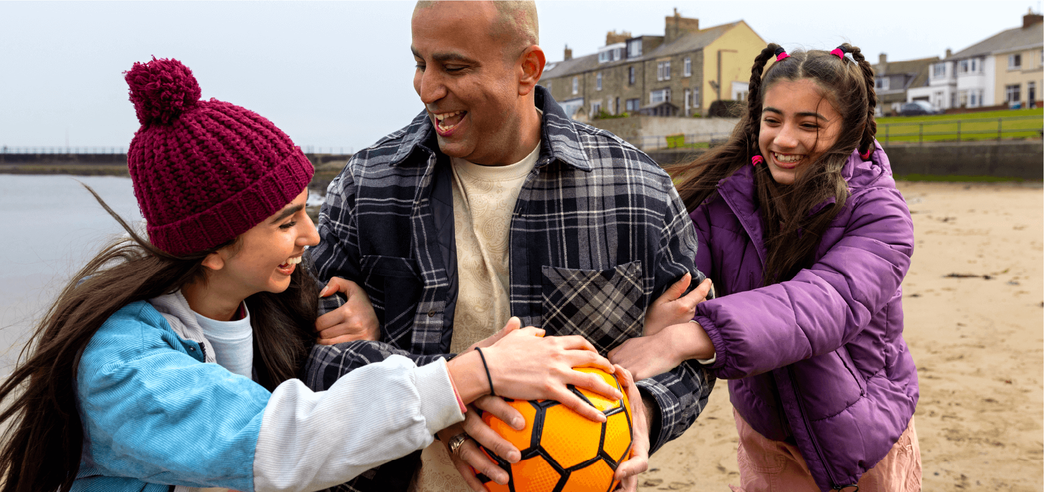 Two children are playfully tugging a football from their father on a sandy beach, with seaside buildings in the background
