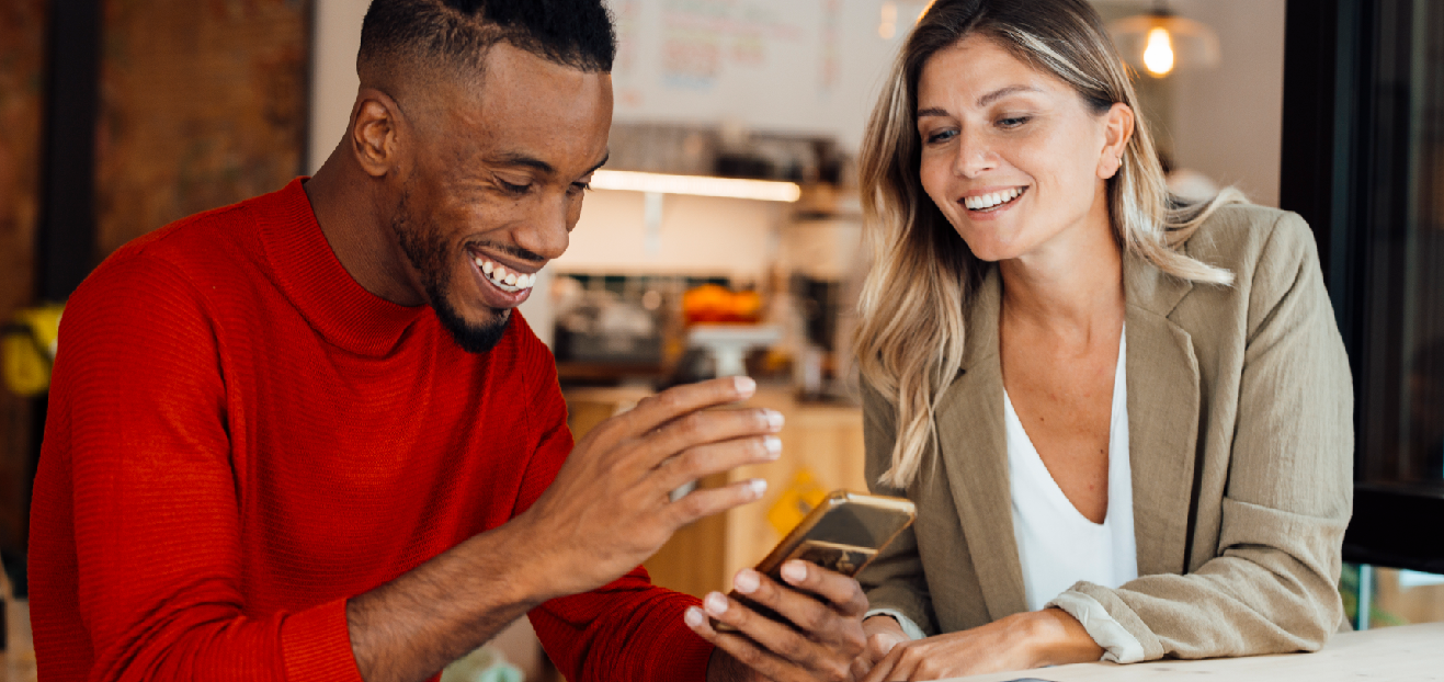 A man in a red sweater gestures animatedly while a woman in a beige blazer looks at a smartphone in a cozy cafe setting
